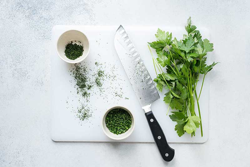 An overhead photo of a white cutting board, chef's knife, and fresh herbs (both chopped and whole). 