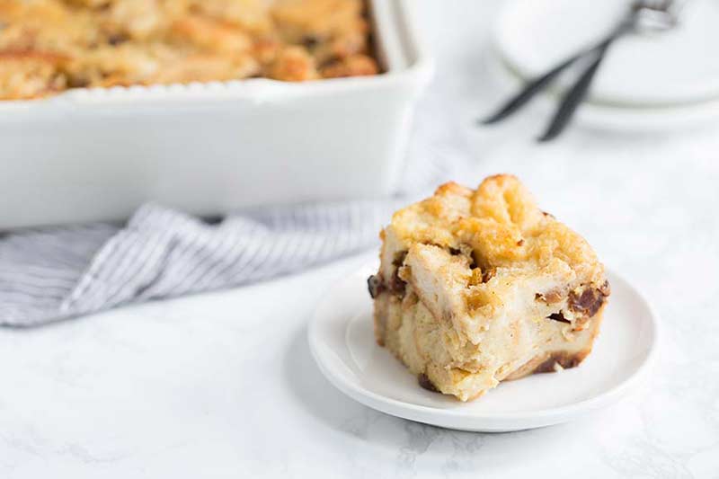 A slice of bread pudding on a plate in the foreground, full bread pudding recipe in the background.