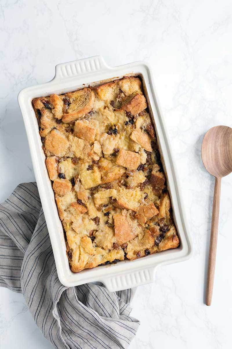 A bread pudding recipe in a white casserole dish, next to a wooden spoon.