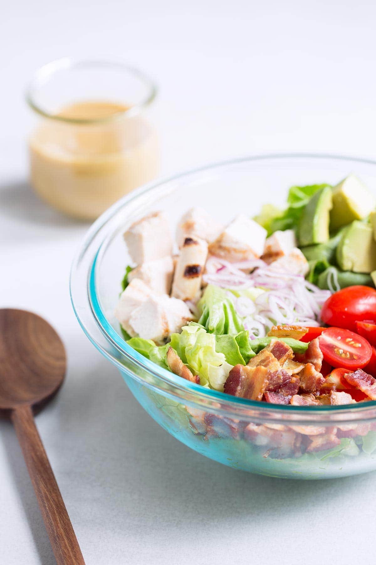 Ingredients for Bacon Avocado Chicken Salad in a large pyrex bowl next to a wood spoon and a jar full of honey mustard.