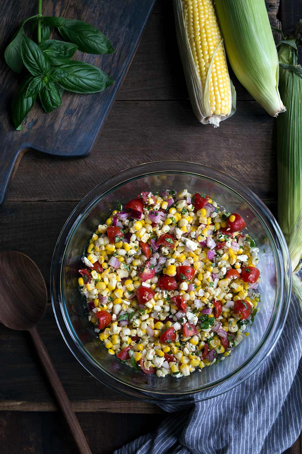 Summer corn salad recipe in a large bowl, surrounded by fresh corn and basil.