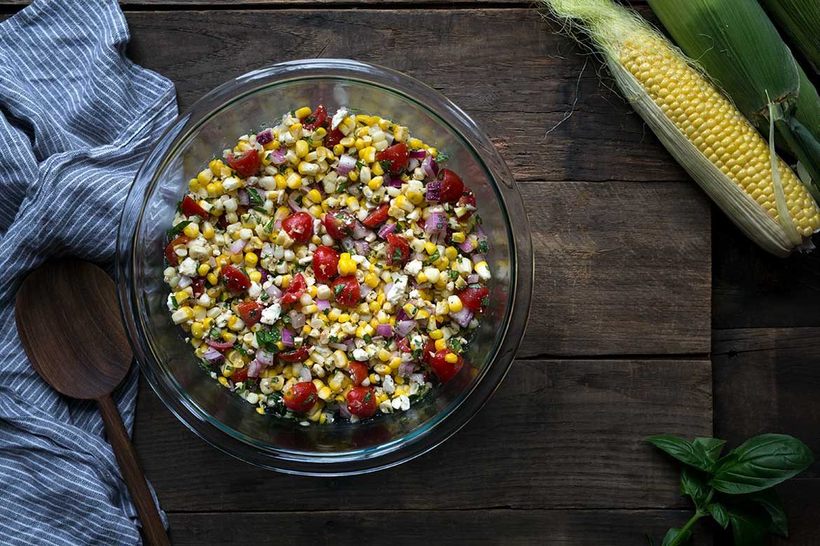 Photo of corn salad recipe in a bowl on a wood backdrop, surrounded by fresh corn and a blue napkin.