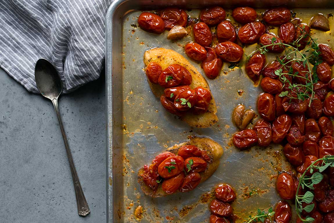 Crostini topped with slow-roasted tomatoes and fresh thyme leaves on a sheet pan, surrounded by more roasted tomatoes and fresh herbs.