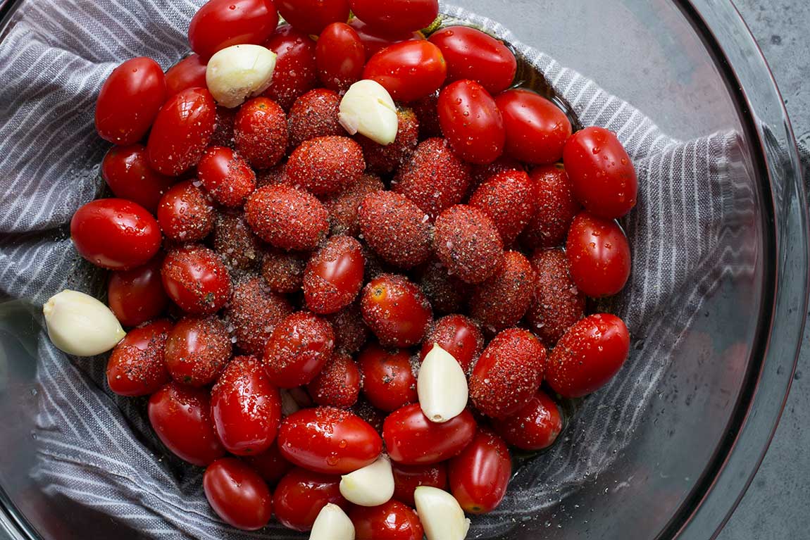 A pyrex bowl containing fresh grape tomatoes, garlic cloves, olive oil, salt, pepper, and sugar.