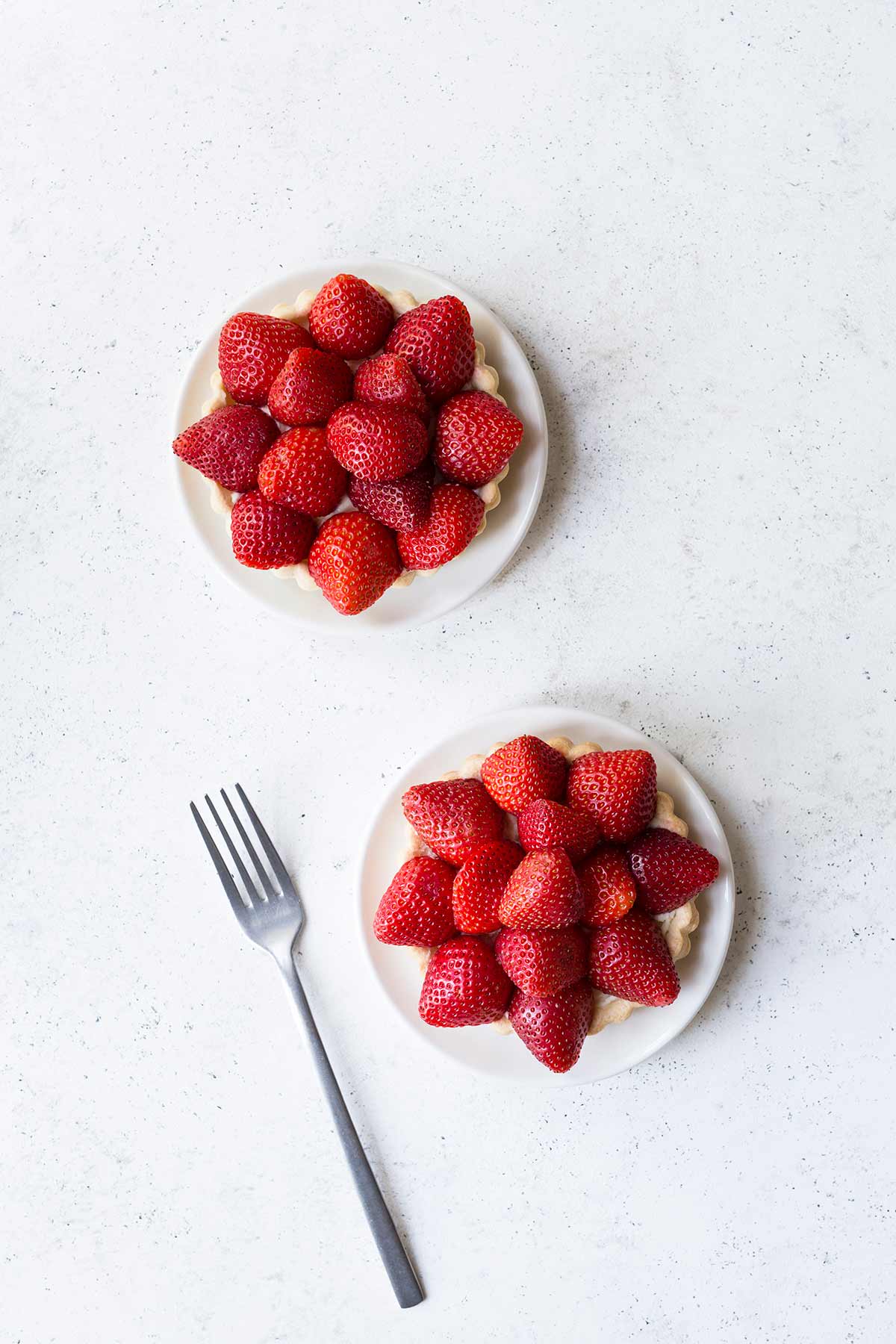 Two strawberry tarts on white plates on a pale backdrop.