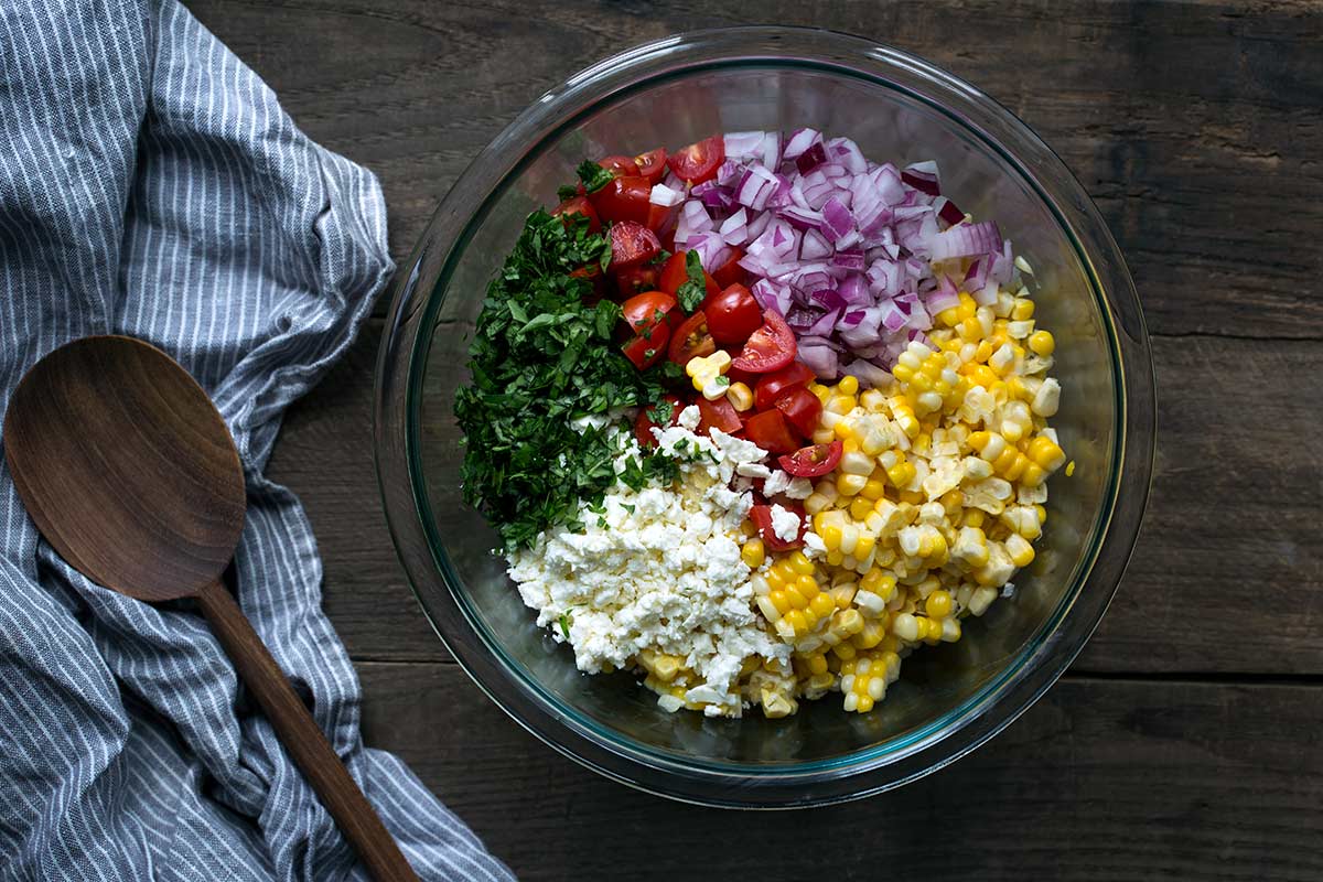 Unmixed ingredients for corn tomato salad in a bowl, including red onions, basil, feta, corn and tomatoes.