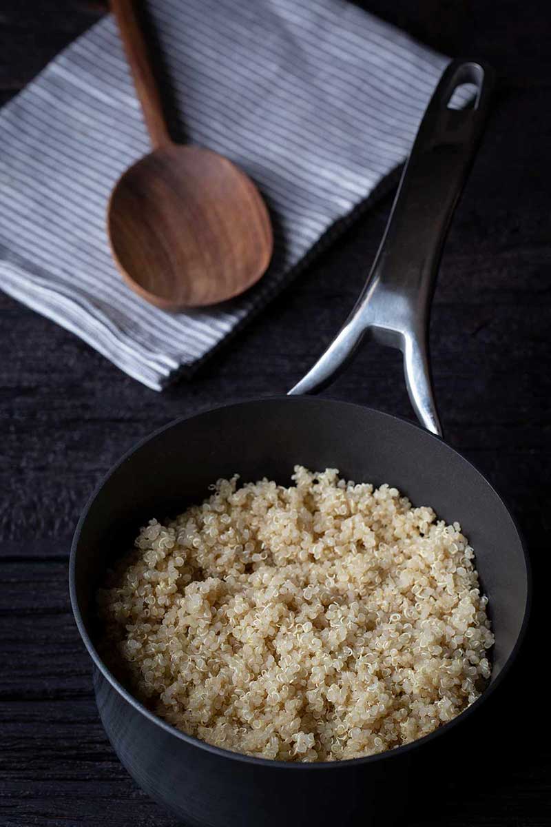 fluffy cooked quinoa in a saucepan with wooden spoon on kitchen towel next to it