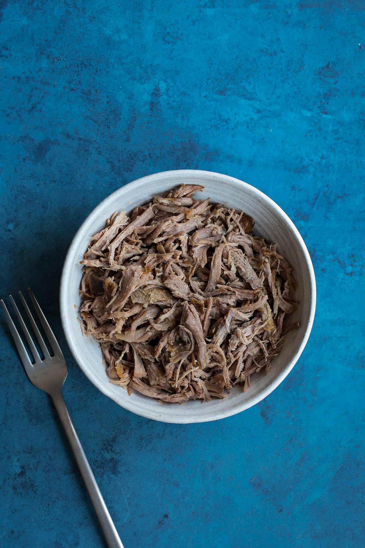 Slow cooker kalua pig in a bowl with a fork on a blue backdrop