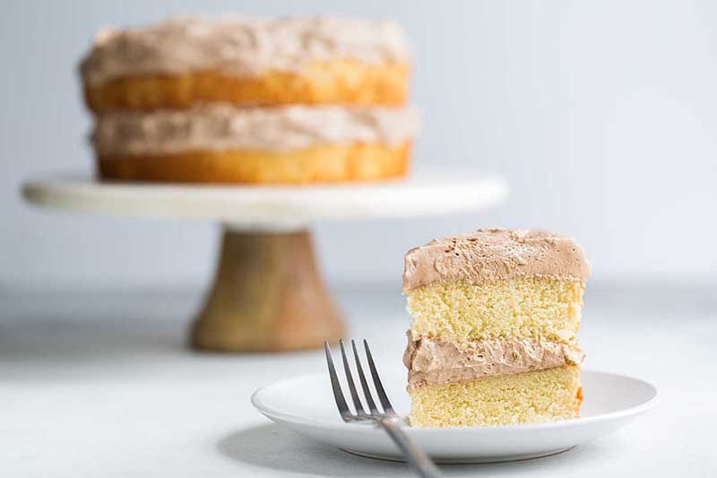 A slice of yellow cake in the foreground. A full double-layer yellow cake with chocolate buttercream is in the background.