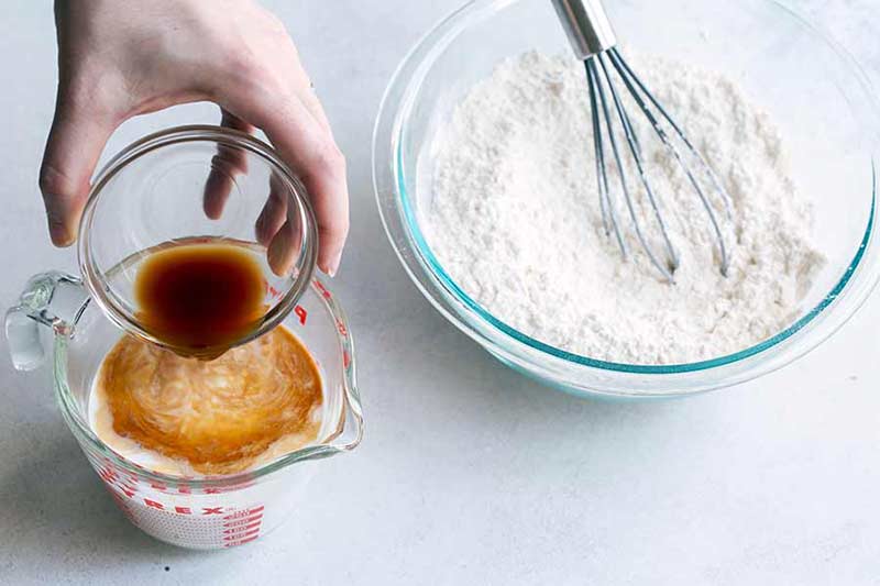 pouring vanilla extract into liquid measuring cup filled with milk, next to glass bowl filled with dry cake ingredients