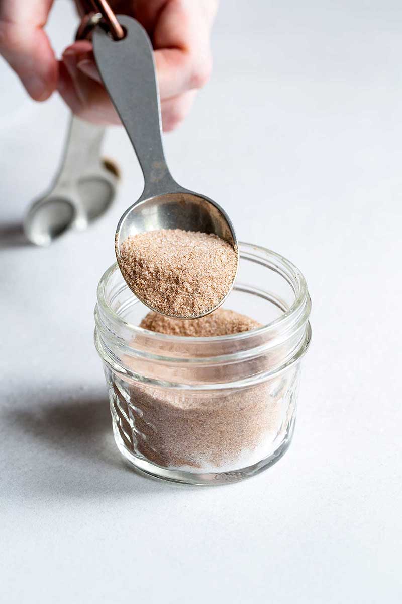 The perfect cinnamon-sugar ratio in a mason jar with a hand holding a measuring spoon full of spice