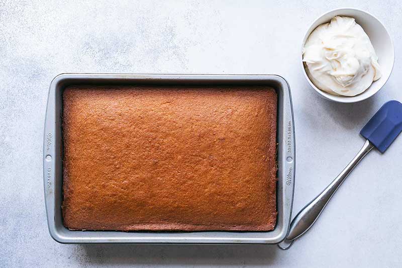 Unfrosted pumpkin cake in pan next to a bowl of cream cheese frosting