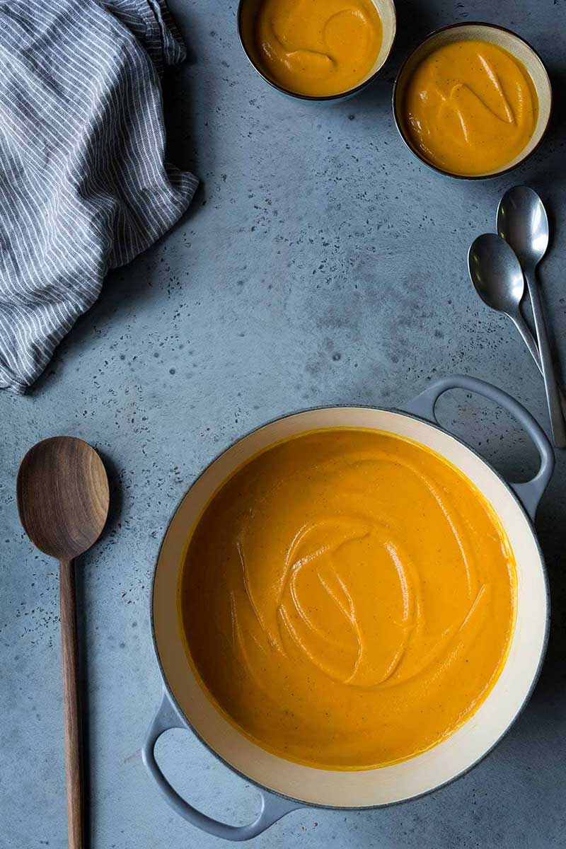 Sweet potato soup in a dutch oven, next to two bowls of soup and a wood spoon.