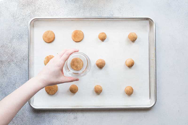 Raw snickerdoodles on a sheet pan, being flattened with a drinking glass.