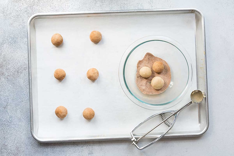 A sheet pan lined with parchment paper, topped with raw snickerdoodles next to a bowl of cinnamon-sugar mix.