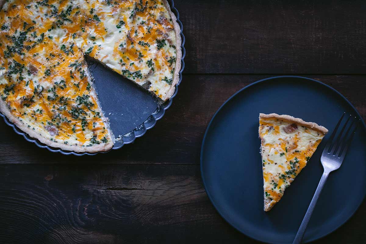 A slice of bacon quiche on a plate next to the remaining quiche.
