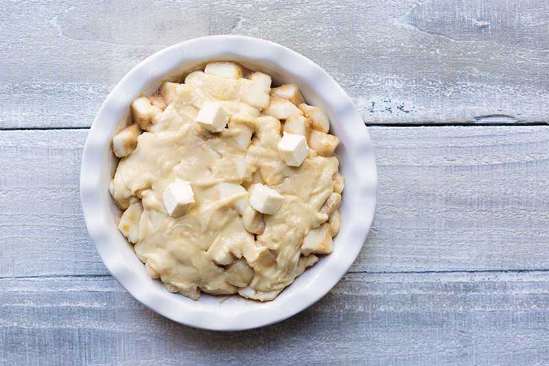 A photo of a pie dish filled with pear filling, topping, and several pats of butter, waiting to be baked.
