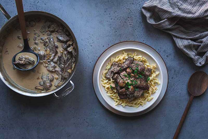 Beef stroganoff in a large flat-sided skillet with a ladle next to a bowl of the recipe served over noodles