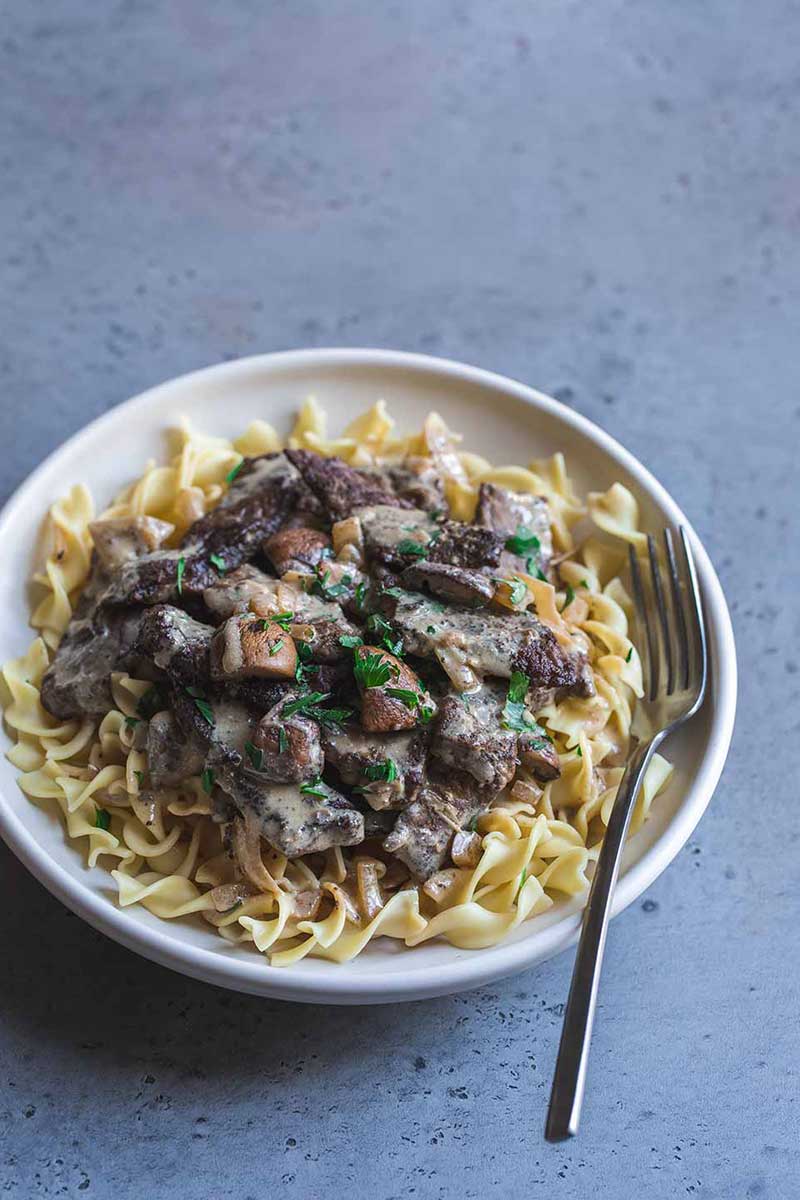 fork on white plate with dinner of beef and mushrooms on bed of buttered egg noodles