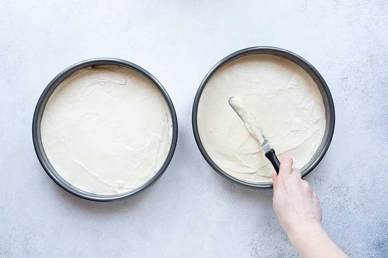 White cake batter being evenly distributed in pans with a spatula