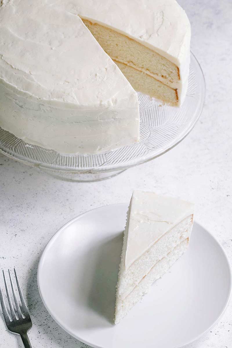 slice of vanilla frosted homemade cake on small white plate with remainder of cake on cake stand behind it