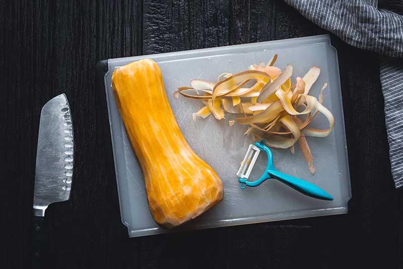 Peeled butternut squash on a cutting board next to a vegetable peeler and the skins