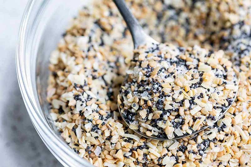 Closeup of everything bagel seasoning ingredients, including minced garlic, minced onion, poppy seeds, and sesame seeds in a bowl with a spoon