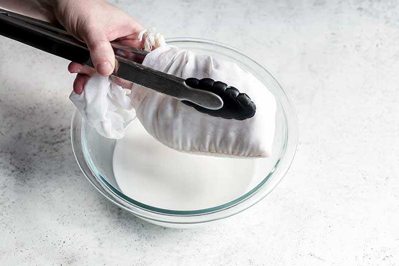 Straining coconut milk through a nut milk bag into a bowl, using kitchen tongs to assist