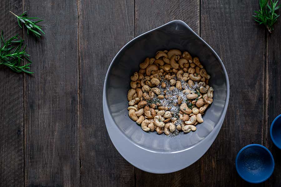 Roasted cashews in a grey bowl with salt, pepper, and rosemary on top prior to mixing