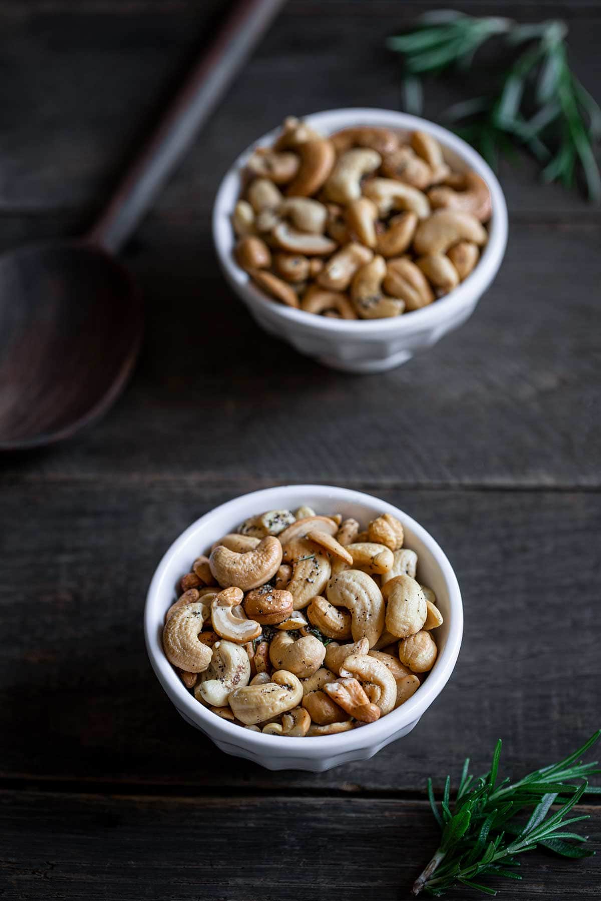 Rosemary salt and pepper cashews in two white bowls next to a wooden spoon
