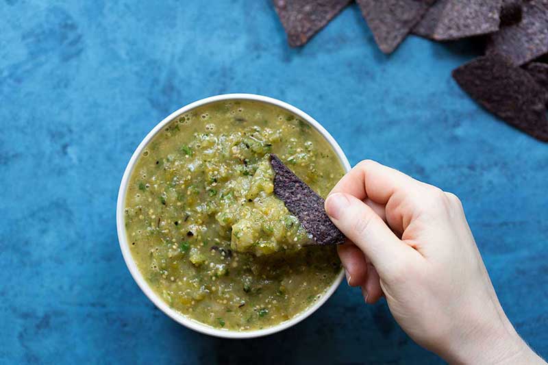 A hand dipping a blue corn chip into a bowl of homemade salsa verde.