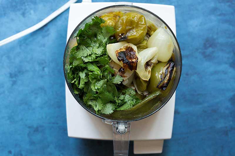 Ingredients for this salsa verde recipe in a food processor, including tomatillos, jalapeños, yellow onions, garlic, lime juice, and cilantro.