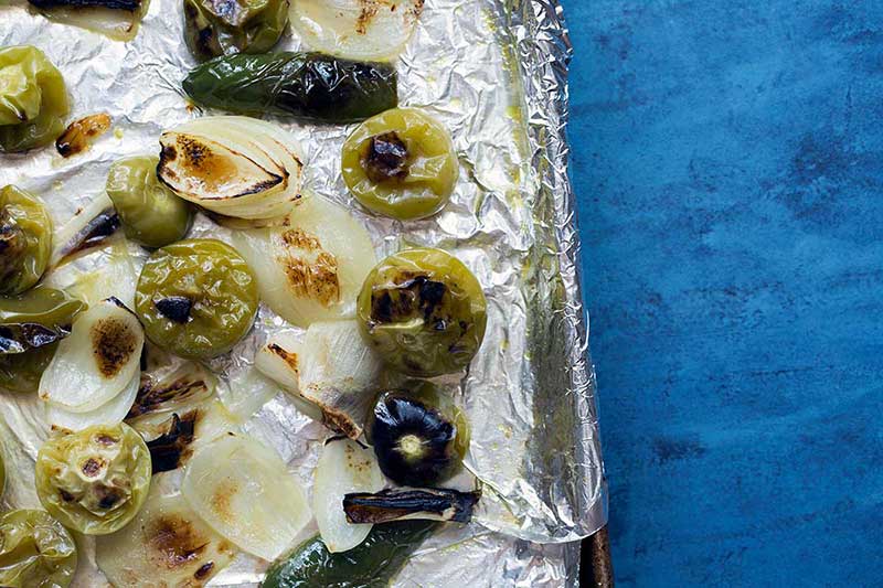 A sheet pan covered with foil, topped with broiled tomatillos, yellow onions, jalapeño peppers, and garlic.