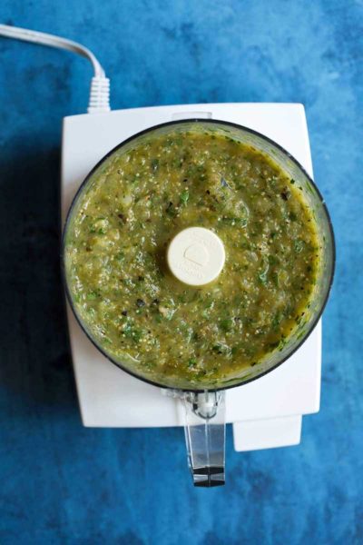 Salsa verde in a food processor on a blue backdrop.