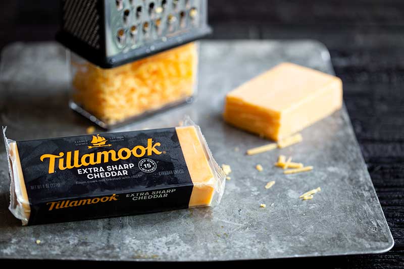 Tillamook extra sharp cheddar in it’s packaging next to a block of cheddar and a box grater filled with cheese.