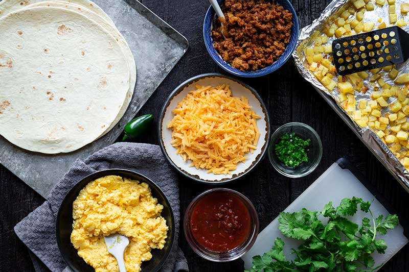 Overhead shot of ingredients laid out for easy breakfast burrito, including cooked chorizo, tortilla craps, cheese, cooked eggs, and salsa.