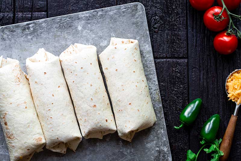 Four rolled breakfast burritos lined up sheet pan