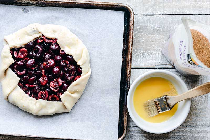 Uncooked galette on a parchment lined sheet pan next to egg wash and turbinado sugar