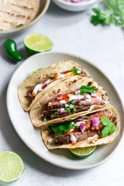 Three grilled flank steak tacos on an oval plate, surrounded by recipe ingredients.