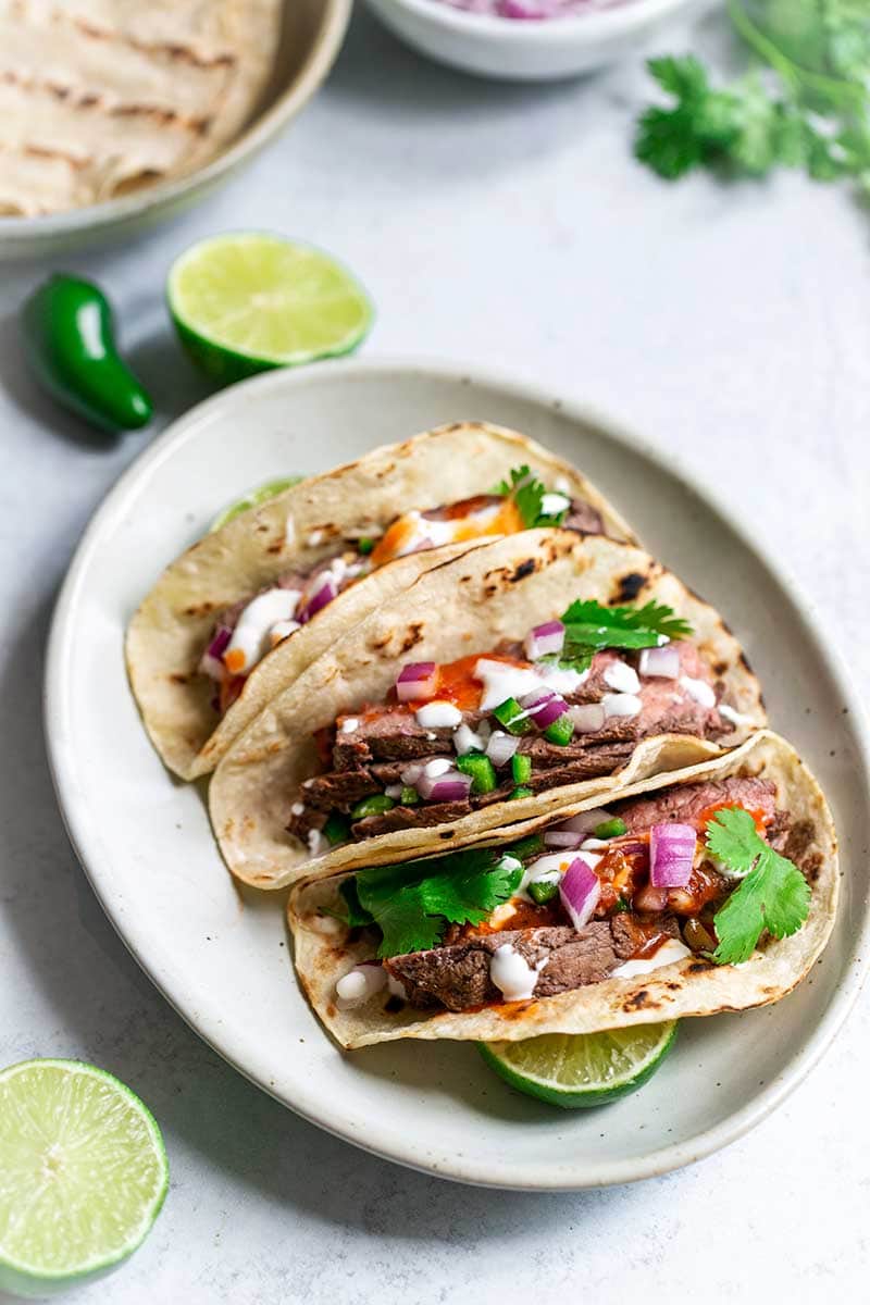 Three grilled flank steak tacos on an oval plate, surrounded by recipe ingredients.