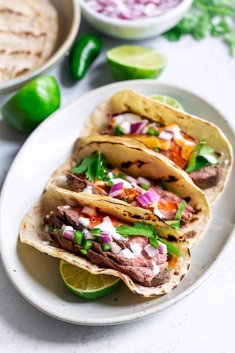 Three grilled steak tacos on an oval plate, surrounded by recipe ingredients.