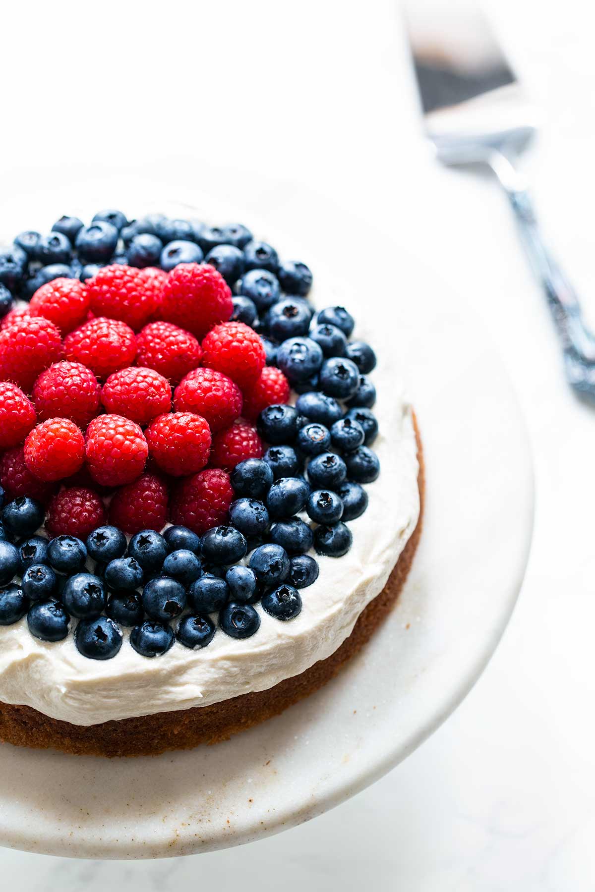 Bright photo of a white cake topped with a thick layer of cream cheese frosting, topped with raspberries in the center and blueberries toward the edge.