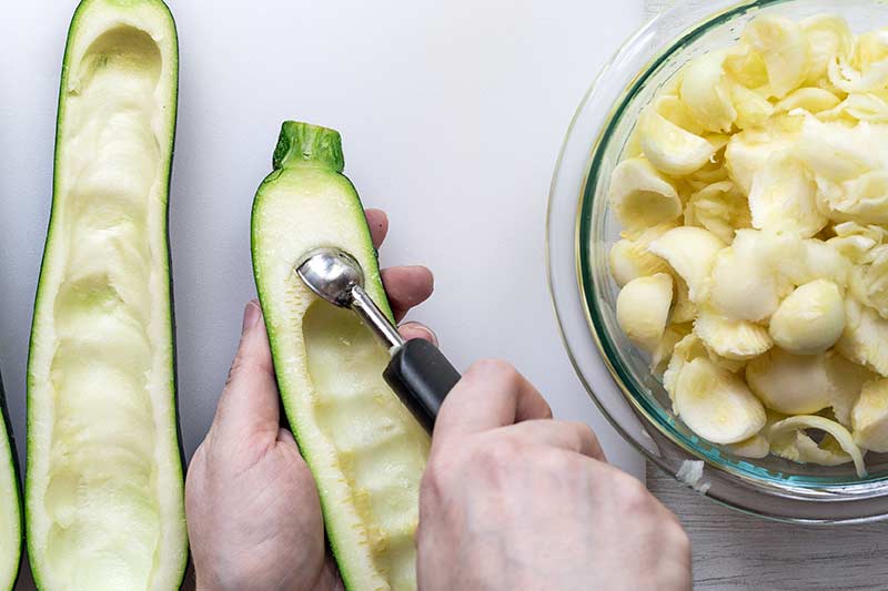Hands using a melon baller to scoop the center seeds out of zucchini halves