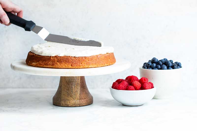 An offset spatula spreading cream cheese frosting over cake. Bowls of raspberries and blueberries sit nearby.