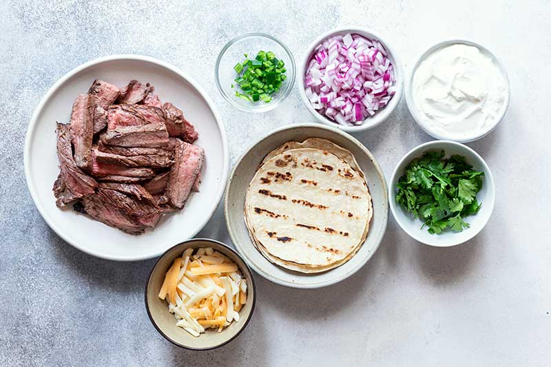 Ingredients for flank steak tacos recipe set out in different size bowls, including sliced steak, grilled corn tortillas, cheese, cilantro, jalapeños, and sour cream. 