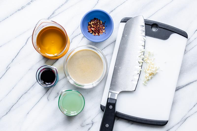 Ingredients for tahini dressing, including tahini, chopped garlic, red pepper flakes, lemon juice, and tamari