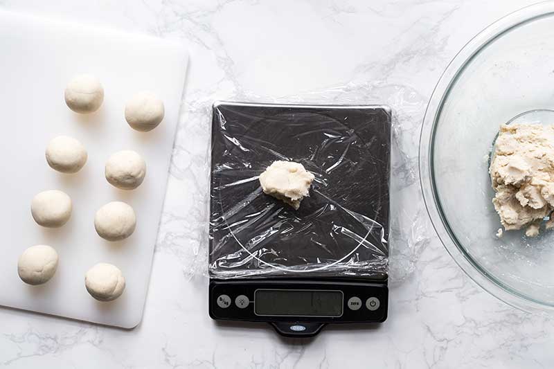 Kitchen scale covered with plastic wrap, topped with a piece of tortilla dough, next to a bowl of dough and a cutting board with portioned dough