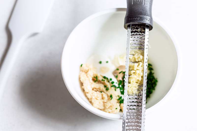 A microplane zester full of minced garlic, set over a bowl of horseradish sauce recipe ingredients