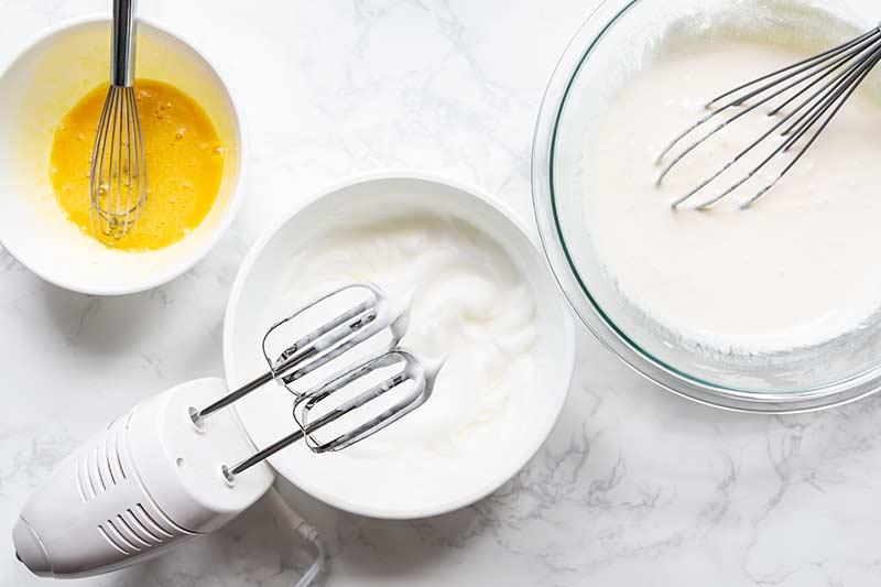 a bowl with whipped egg whites, a hand mixer, a bowl with whisked egg yolks, and a bowl with the remaining souffle ingredients