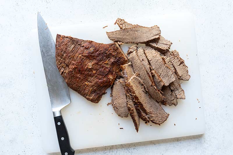 Sliced braised beef on a cutting board next to a slicer/carving knife.